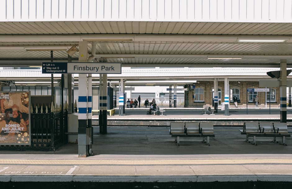 A view of Finsbury Park train station platform with several passengers waiting, framed by a white and blue sign displaying the station's name. The platform features metal benches, some occupied by com