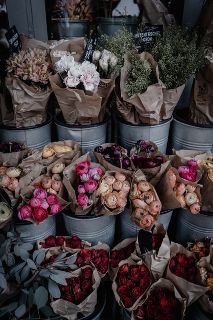 An outdoor display of various fresh flowers arranged in metal buckets and wrapped in brown paper, including roses in shades of peach, pink, and deep red, along with white carnations, grey eucalyptus l
