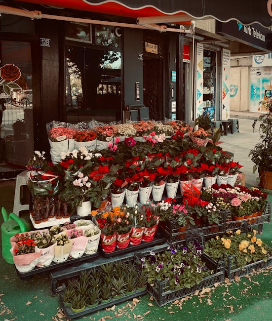 A vibrant outdoor display of fresh flowers in front of a shop on a sidewalk, featuring a variety of blooms including pink and white roses, red tulips, orange and yellow marigolds, purple pansies, and 