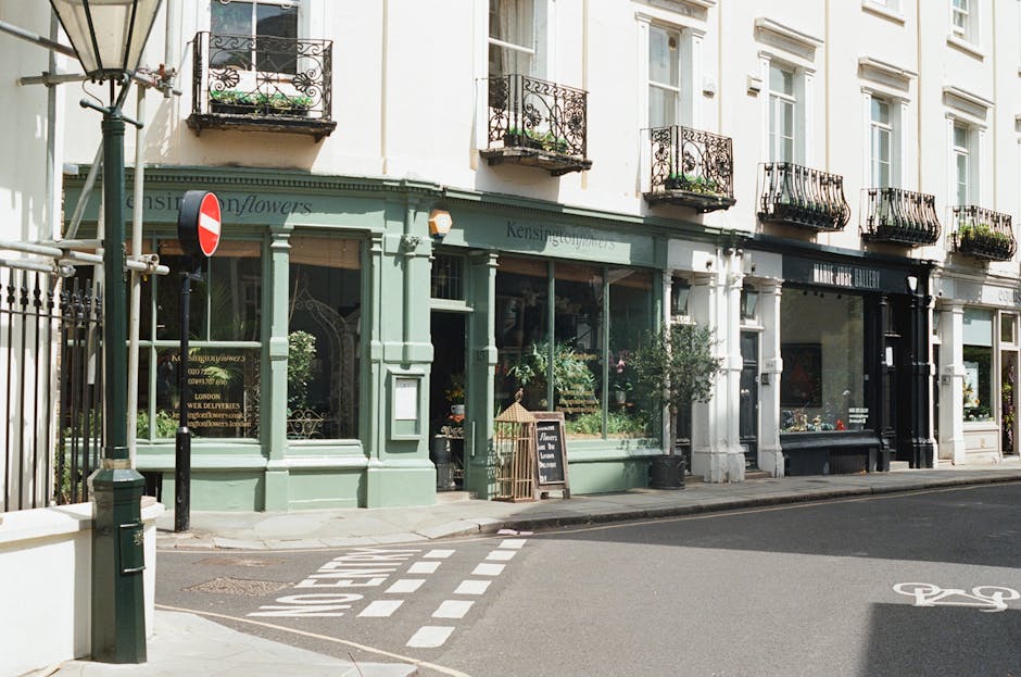 A storefront of a flower shop on a quiet street corner in Stroud Green, N4, featuring a light green painted facade with large glass windows displaying various floral arrangements. The shop has decorat