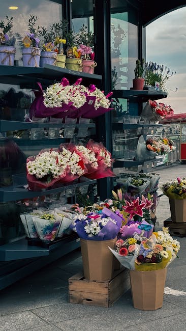 A display of various floral arrangements on shelves and in buckets at a flower shop, showcasing fresh flowers including white daisies, pink roses, and purple statice, wrapped in colorful paper and pla
