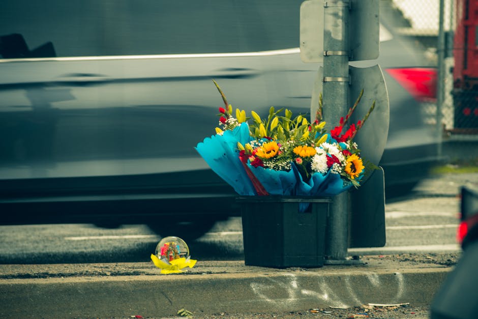 A black plastic delivery box filled with a colorful bouquet of fresh flowers, including yellow sunflowers, red and white daisies, and greenery, is placed on the pavement next to a metal pole on a city