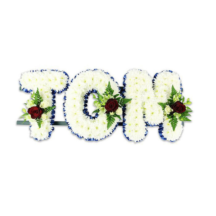 A poignant and meticulously made memorial arrangement spells TOM in large, rounded letters formed from countless small, immaculate white chrysanthemums. The chrysanthemums are packed tightly to create a plush, sculpted surface that appears almost cloud-like; their delicate petals catch light across the face of each letter, giving depth and a soft tactile impression. The perimeter of each letter is finished with a narrow royal blue ribbon that creates a crisp, dignified border and visually separates the white flowers from the background. Inside each letter sits an arranged floral spray: a single deep red rose anchors each cluster, evoking love and remembrance, while clusters of small cream-toned spray roses and graceful fern fronds provide contrast, texture and a gentle green frame. There is a quiet, restrained palette - white, royal blue, deep red and fresh green - that supports the arrangement's solemn purpose. A slender dark green or black metal support bar is visible beneath the composition, suggesting it is designed to be displayed upright on a stand at a service. The tribute is presented against a stark white backdrop and lit with soft, even light to emphasize the sculptural form and the vividness of the blue trim and red focal roses. This classic style of bespoke name tribute is commonly chosen for funerals and memorial services when families want a personalised, handcrafted expression of sympathy, carefully prepared by experienced florists for dignified presentation at churches, crematoria or community chapels around Finsbury Park, Manor House and nearby neighbourhoods.