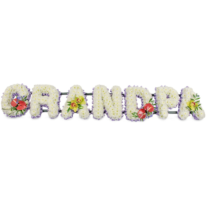 This image shows a long, horizontal floral arrangement spelling out "GRANDPA" in large, three-dimensional capital letters, meticulously handcrafted for a funeral or memorial. Each letter is made from masses of small, white chrysanthemums, packed so closely that the flowers merge into a smooth, plush surface, suggesting purity, serenity, and a comforting presence. The clean white background allows the tribute to stand alone, as though it has just been gently placed on the floor of a Finsbury Park chapel or in the flower room of a nearby funeral director. At the base of the letters, a soft border of pale lavender ribbon adds a delicate band of colour, edging the white blooms with a cool, almost silver-lilac outline that enhances the clarity of each shape. Supporting bars in dark green run behind and beneath the letters, barely visible yet essential, giving structure so that the tribute can be moved from a florist's studio to a service with ease. Vivid accent sprays punctuate the design: the 'G' and 'D' are each embellished with clusters of deep red anthuriums, their waxy, heart-shaped petals and bright yellow spadices framed by glossy green foliage and touches of smaller filler flowers. These strong crimson notes bring warmth and intensity to the tribute. The first and last 'A' are softened with sprays of yellow-green orchids, their elegant, arching stems and sculpted blooms suggesting admiration and enduring love. Altogether, the tribute feels bespoke and deeply personal, like a custom piece ordered to honour a cherished grandfather from the Finsbury Park area, expressing gratitude, remembrance, and respect in a clear, floral script.